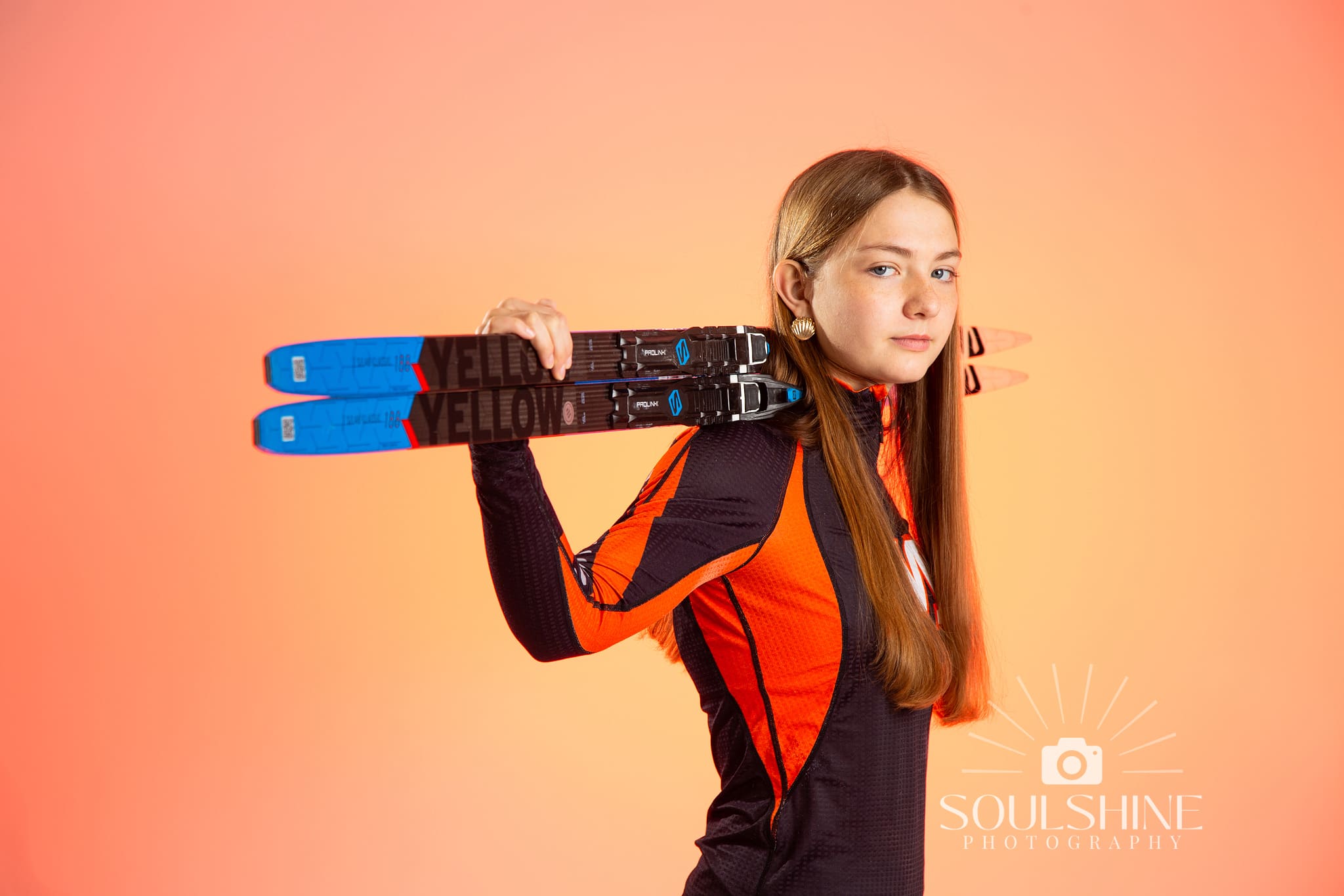 Girl holding nordic skis on her shoulders. Studio image with orange background.
